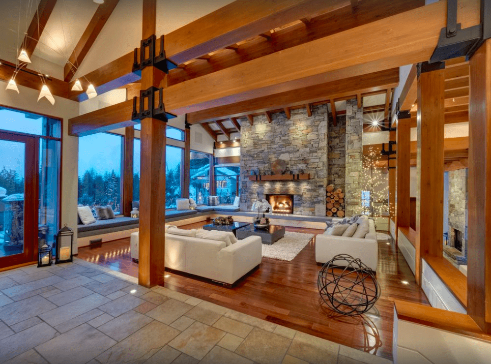 Photo of the Belmont Estate great room, with wooden beams, couches, fireplace with stone accents and a view outside of the snow in the evening.